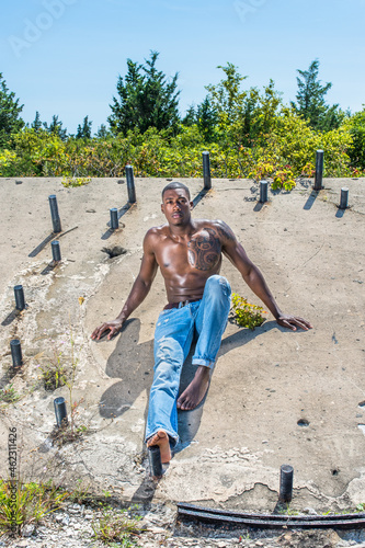 Obraz na płótnie A masculine black guy, wearing blue jeans, barefoot and half naked, is sitting a old construction site, relaxing.. w salonie