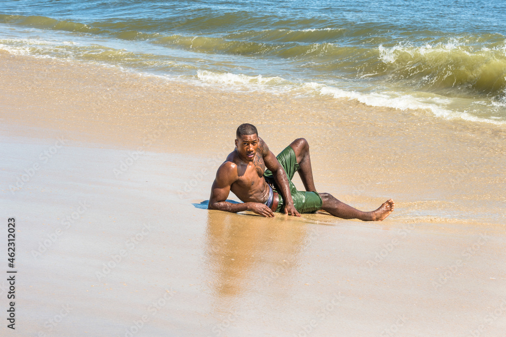 A well built young black guy, half naked, is lying on the beach and playing with waves of water..