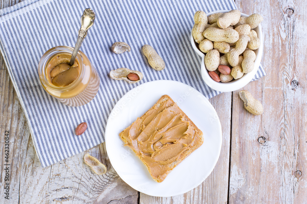 Peanut butter in a glass jar and a piece of bread with spread paste on a wooden background. Delicious and healthy breakfast. Peanut butter in a glass jar and a piece of bread with spread paste on a wooden background. Delicious and healthy breakfast.