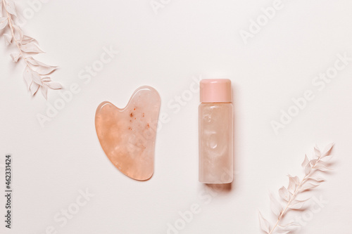 Guashe scrapes from rose quartz and moisturizing gel on pale pink background, decorated white dry flowers. Natural stone massager for face photo