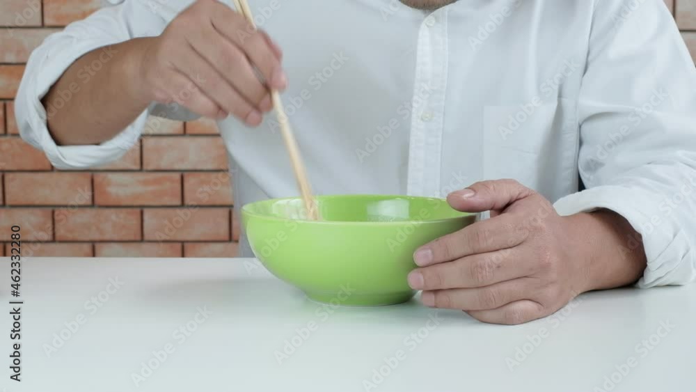 Hungry Thai man in white shirt uses chopsticks to eat hot instant noodles in green bowl in lunch breaks, quick, tasty, and cheap. Traditional Asian fast food meal of Japanese and Chinese lifestyle. Hungry Thai man in white shirt uses chopsticks to eat hot instant noodles in green bowl in lunch breaks, quick, tasty, and cheap. Traditional Asian fast food meal of Japanese and Chinese lifestyle.