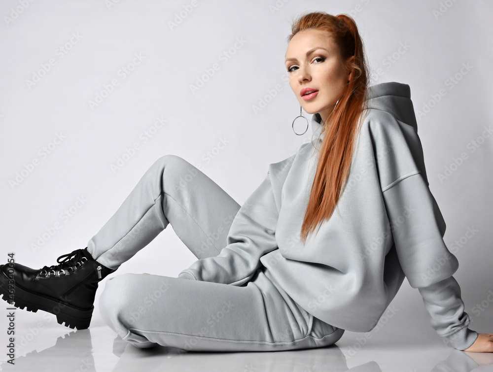 Fashionable young sexy woman wearing sportswear hoodie, pants and black massive  shoes sits on floor side to camera, demonstrating her long straight healthy  red hair and looking at camera Photos | Adobe