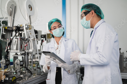 Both male and female Asian scientists are outfitted with protective gear. Examining the factory's machinery and materials used in the mask-making process. Covid 19 is a piece of preventive equipment.