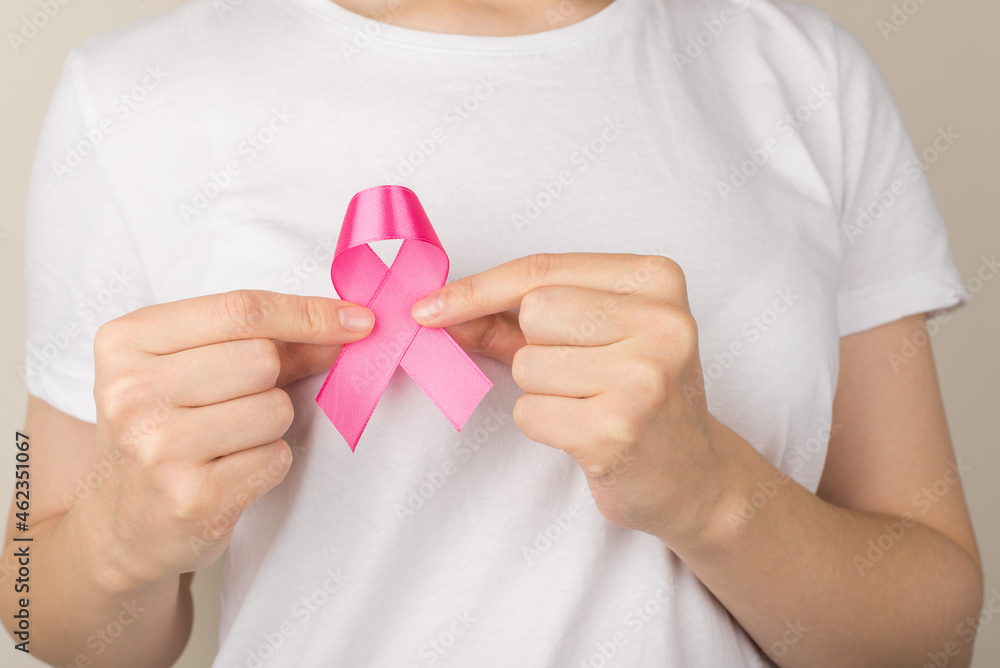 Close up photo of girl in white t-shirt holding pink ribbon symbol of breast cancer awareness on isolated grey background Close up photo of girl in white t-shirt holding pink ribbon symbol of breast cancer awareness on isolated grey background