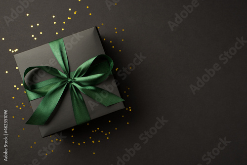 Top view photo of stylish giftbox with green ribbon bow and golden confetti on isolated black background with empty space