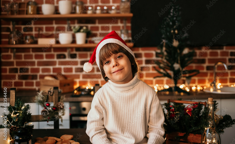 portrait of a little boy in a white sweater and a red Christmas hat sitting on a table in the kitchen with