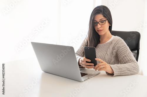Young woman with glasses working in office using smartphone and laptop in backlight