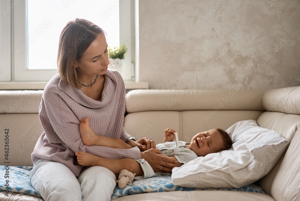 Worried young mother sitting on sofa beside her sick son with high fever. Mom measures temperature using thermometer of sick child lying under blanket at home. Worried young mother sitting on sofa beside her sick son with high fever. Mom measures temperature using thermometer of sick child lying under blanket at home.