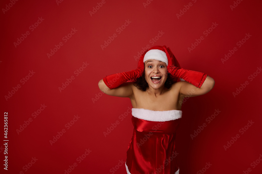 Beautiful woman in Santa carnival costume holds her hands on the head and rejoices looking at camera, expressing surprise, isolated over red background with copy space for Christmas and New Year ad Beautiful woman in Santa carnival costume holds her hands on the head and rejoices looking at camera, expressing surprise, isolated over red background with copy space for Christmas and New Year ad