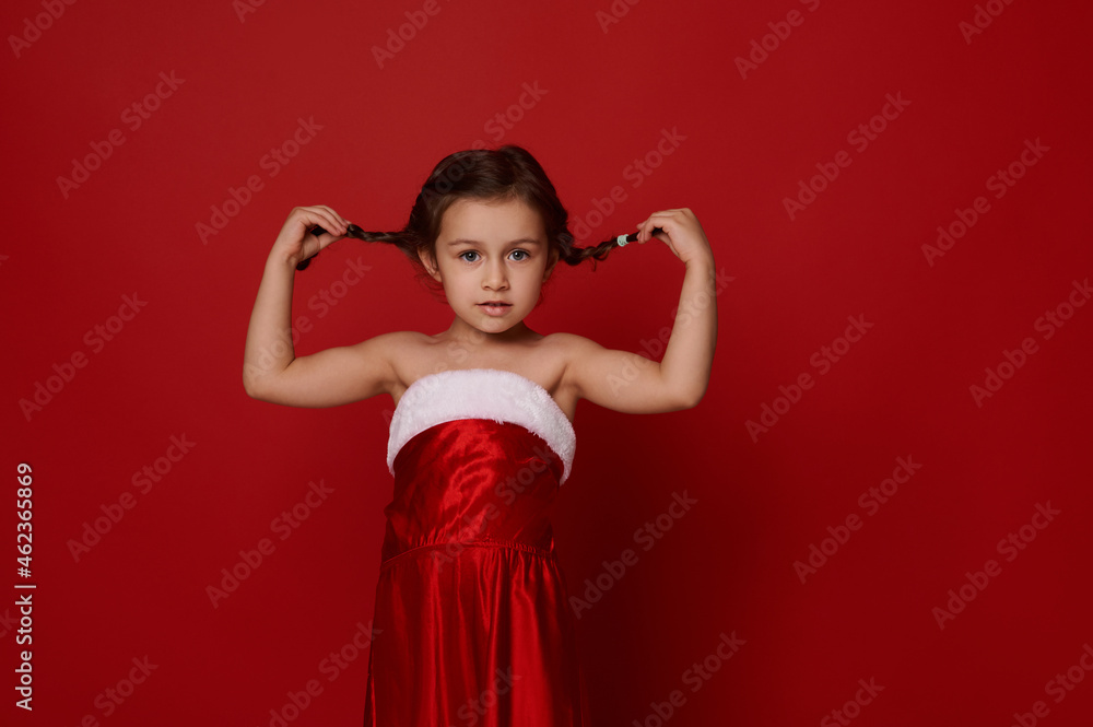 Adorable pretty 4 years old little girl, cute child in Santa Claus clothes poses holding pigtails,looks at camera. Isolated over red background with copy space for Christmas and New Year advertising Adorable pretty 4 years old little girl, cute child in Santa Claus clothes poses holding pigtails,looks at camera. Isolated over red background with copy space for Christmas and New Year advertising
