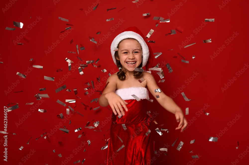 Cheerful little girl, adorable child dressed in Santa carnival costume rejoices, smiles toothy smile , throwing confetti and sequins over red background with copy space. Christmas, New Year concept