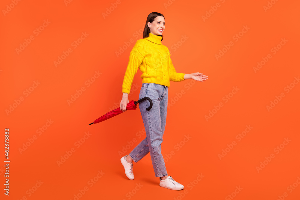 Full size profile side photo of young woman happy positive smile go walk wet forecast umbrella isolated over orange color background Full size profile side photo of young woman happy positive smile go walk wet forecast umbrella isolated over orange color background
