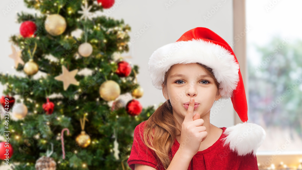 winter holidays and children concept - happy girl child in santa helper hat making shh gesture over christmas tree background