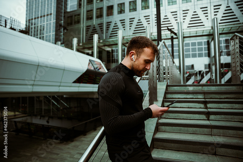 Caucasian male athlete leaning against bridge scrolling on cellular device lsitening t o music with earphones