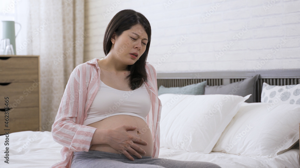 pregnant female is leaning backward to support herself with hand on bed while feeling strong contraction and groaning in pain at home. pregnant female is leaning backward to support herself with hand on bed while feeling strong contraction and groaning in pain at home.
