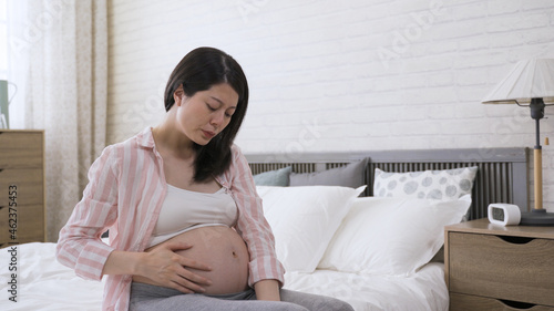 Photos portrait asian woman in her last trimester is groaning and holding her belly while experiencing contraction pain by bedside in the bedroom