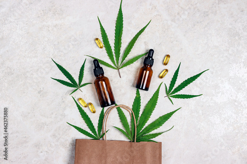 Brown paper shopping bag with green marijuana leaves, Cannabis extract oil in a bottles and capsules on light marble background.