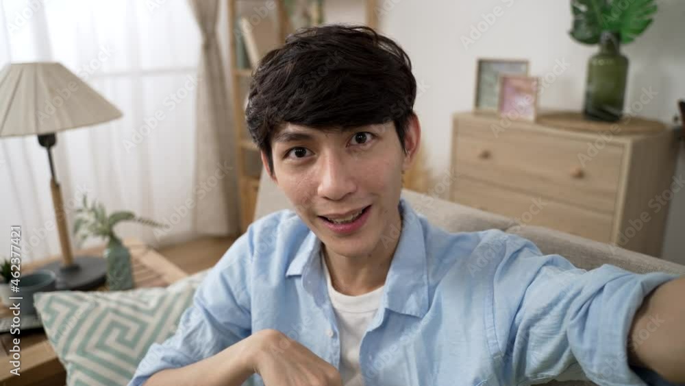 mobile cam view of cheerful asian guy waving hand and finger pointing at camera while having pleasant talk with friend via video call in the living room at home mobile cam view of cheerful asian guy waving hand and finger pointing at camera while having pleasant talk with friend via video call in the living room at home