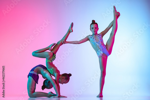 Fototapeta Naklejka Na Ścianę i Meble -  Three little beautiful rhythmic gymnastics artists, girls in sport event isolated on gradient pink blue studio background in neon light