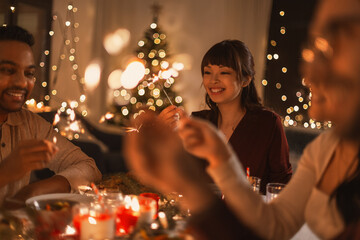 holidays and celebration concept - multiethnic group of happy friends with sparklers having christmas dinner at home