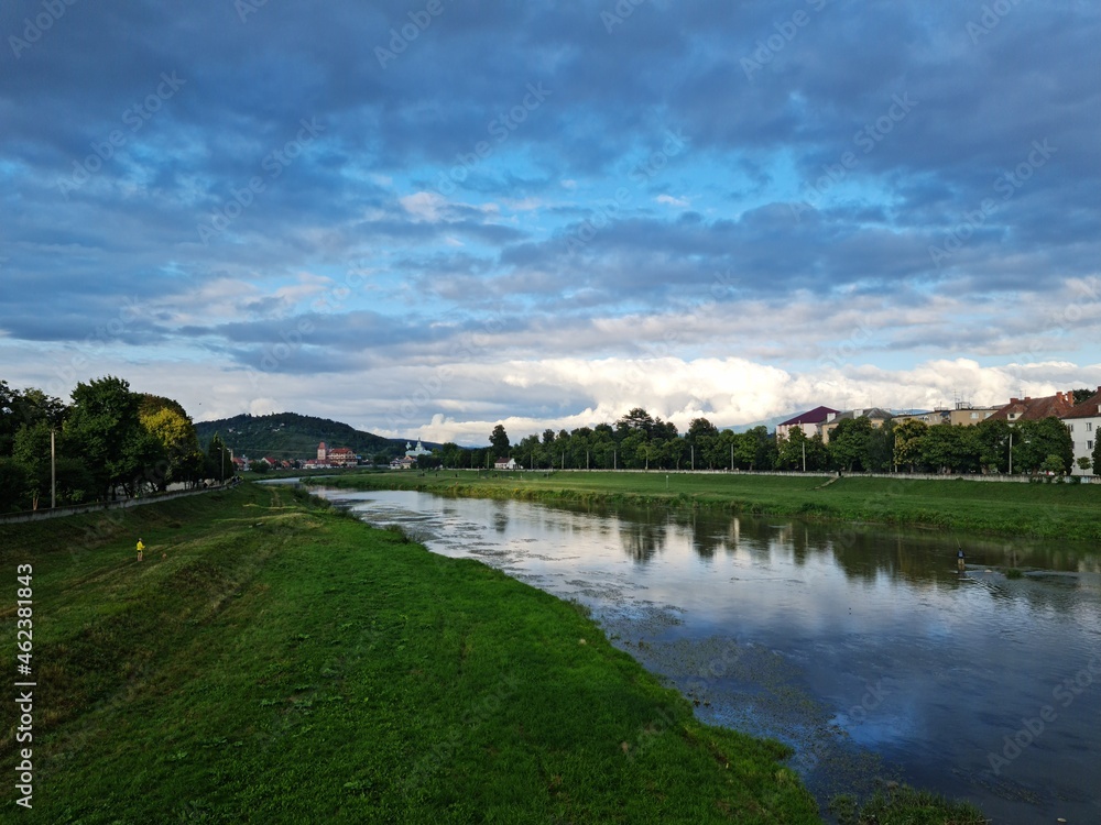 Natural beauty of blue sky and calm water of river in Mukachevo, Ukraine. Beautiful landscape with river and houses at sunset. Scenic view of the old town embankment on a cloudy summer day at dusk.