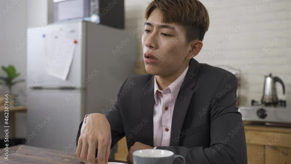 closeup depressed Asian male in business suit is lack of energy while eating bread for breakfast before work in the dining room at home. closeup depressed Asian male in business suit is lack of energy while eating bread for breakfast before work in the dining room at home.