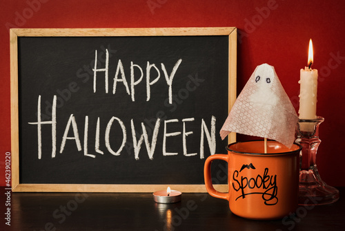 happy halloween lettering on chalk board, Cup with drink next to burning candles and halloween pumpkin on bloody background