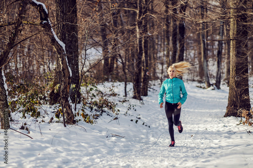Fit sportswoman jogging on snowy path in forest at winter. Healthy lifestyle, winter fitness