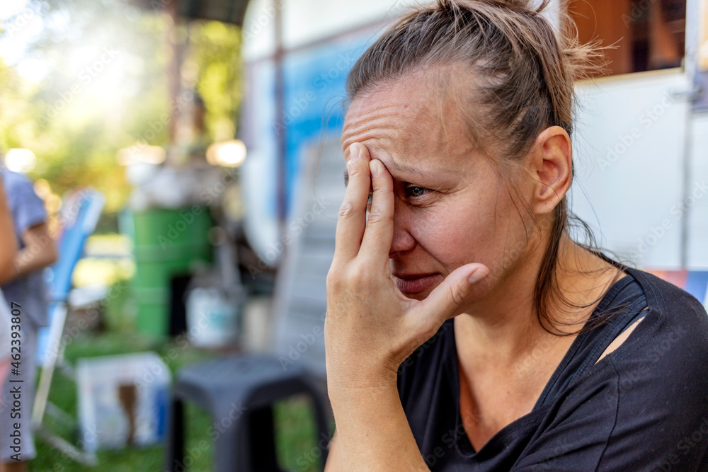 Stressed depressed young mother sits outside in a playground with her kids playing behind her. Single frustrated woman hold her head with hands sitting on chair with playful kids on a background. Stressed depressed young mother sits outside in a playground with her kids playing behind her. Single frustrated woman hold her head with hands sitting on chair with playful kids on a background.