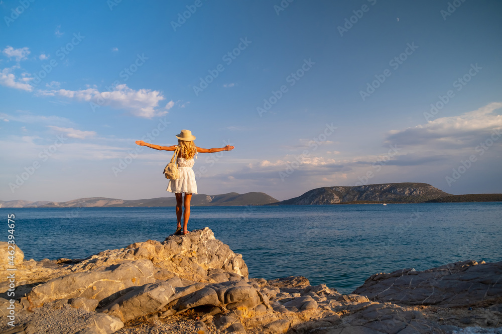Beautiful young woman wearing traw hat standing in rocks in white dress by the seashore Beautiful young woman wearing traw hat standing in rocks in white dress by the seashore