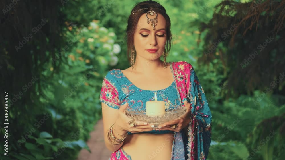 Fantasy Indian woman princess holding crystal candlestick, candle burning bright flame. Beautiful girl blue pink national vintage sari dress. Shawl silver tiara on head. Summer garden rainforest India Fantasy Indian woman princess holding crystal candlestick, candle burning bright flame. Beautiful girl blue pink national vintage sari dress. Shawl silver tiara on head. Summer garden rainforest India