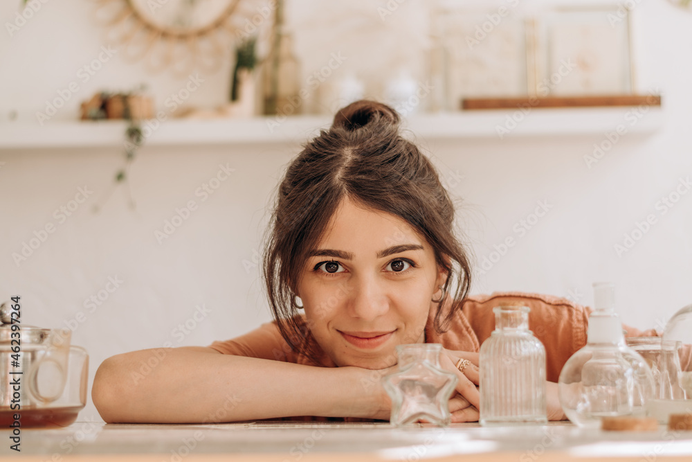 Portrait of a beautiful young woman of mixed race posing at a table on which there are glass vases and bottles.In the background there are shelves with indoor plants and decorative elements.