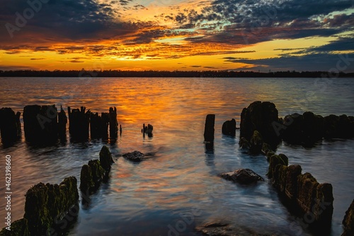 Beautiful sunset in the sky with clouds over the river. Lines of old wooden supports from the pier in the water in the foreground. Landscape at sunset