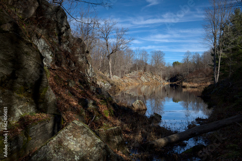 Cliff and Pond in Autumn - Frontenac Provincial Park, Ontario Canada. A view past a rocky cliff towards a serene pond reflecting wispy clouds and blue sky.