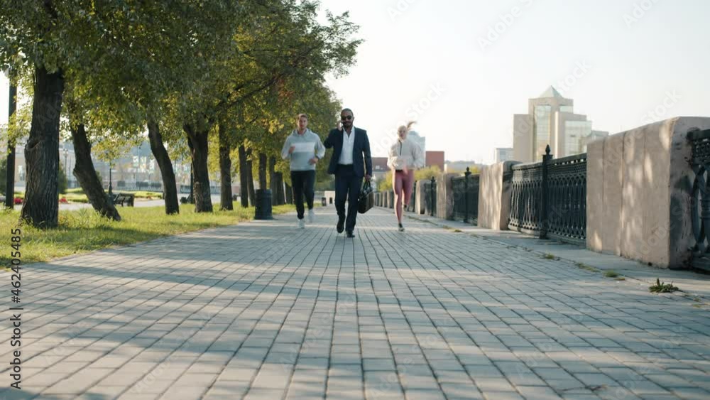 Businessman is walking and talking on mobile phone while sports people are jogging in city street enjoying healthy activity outdoors Businessman is walking and talking on mobile phone while sports people are jogging in city street enjoying healthy activity outdoors