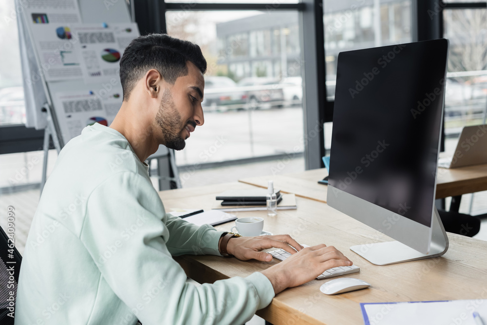 Fototapeta premium Side view of smiling arabian businessman using computer near coffee in office