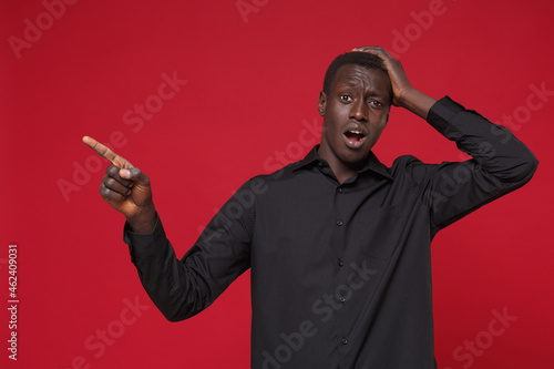Shocked young african american man in classic black shirt posing isolated on red background in studio. People lifestyle concept. Mock up copy space. Pointing index finger aside up, put hand on head.