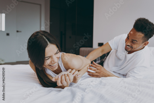 Vivid young couple two family in casual white clothes lying in bed on stomach man tickling woman fooling around time together in bedroom lounge home house wake up dream be lost in reverie good day photo
