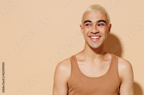 Young smiling laughing cheerful blond latin american gay man 20s with make up in beige tank shirt looking aside isolated on plain light ocher background studio portrait People lgbt lifestyle concept photo