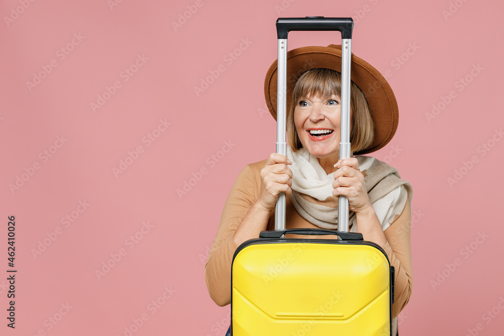 Traveler tourist fun mature elderly senior woman 55 years old wear brown shirt hat scarf hold suitcase bag hide behind handle look aside isolated on plain pastel light pink background studio portrait Traveler tourist fun mature elderly senior woman 55 years old wear brown shirt hat scarf hold suitcase bag hide behind handle look aside isolated on plain pastel light pink background studio portrait