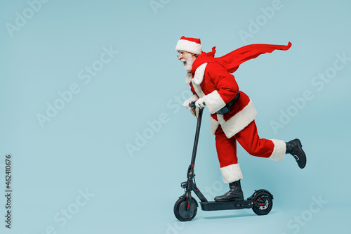 Full body side view fun cool old Santa Claus man 50s in Christmas hat red suit super hero coat rides kick scooter isolated on plain blue background studio. Happy New Year 2022 merry ho x-mas concept,