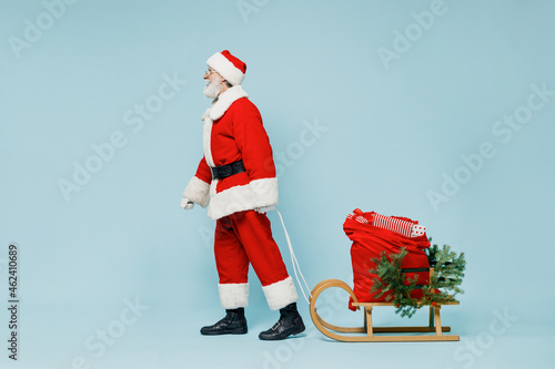 Full body old Santa Claus man 50s in Christmas hat red suit clothes go with gifts sled walking isolated on plain blue background studio. Happy New Year 2022 celebration merry ho x-mas holiday concept.