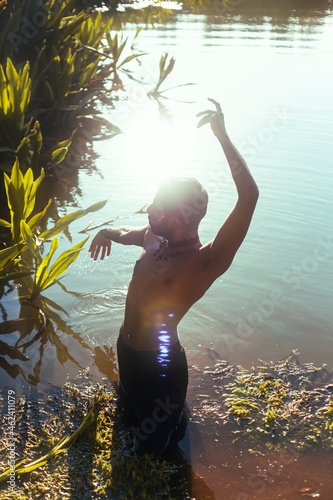 portrait of a dark skinned Indian man by a lake, shirtless, with make up, surrounded by nature photo