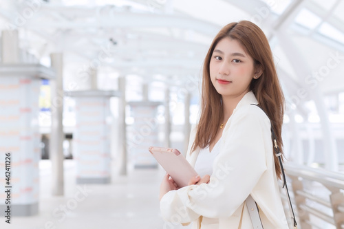 Free Download Beautiful young age Asian professional working business woman in white suit is outdoors to meet a client at the office her holding hand tablet with business buildings and city in background. - GFXLoad