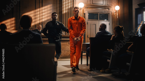 Cinematic Court of Law and Justice Trial Proceedings: Portrait of Accused Sad Male Criminal in Orange Jumpsuit Led Away by Security Guard in Front of Judge and Jury. Sentenced to Serve Jail Time.