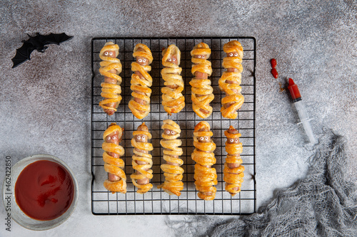 Halloween mummy hot dogs. Sausages with eyes wrapped in dough on cooling rack on concrete background. Crazy funny Halloween food. Top view, flat lay.