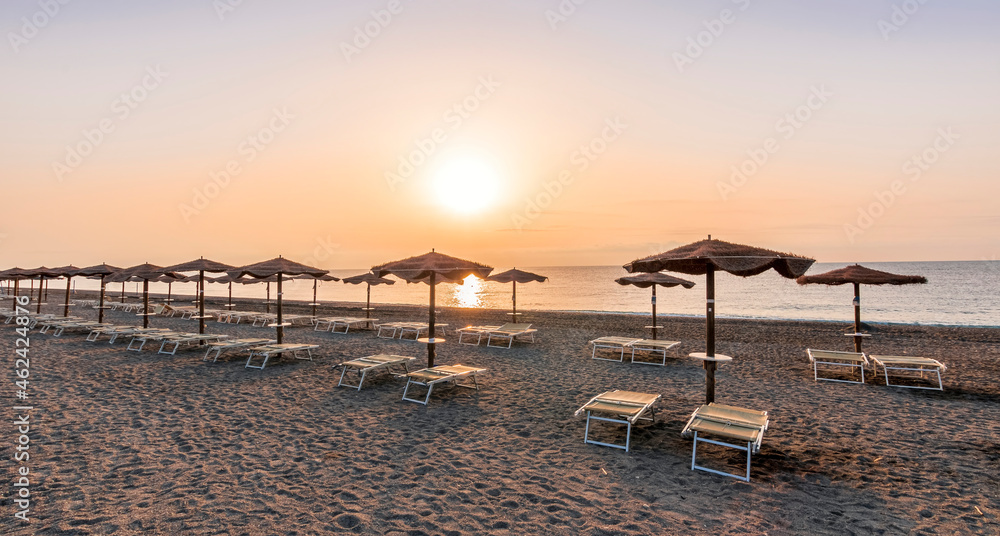 scenic view at nice beach with rows of umbrellas and chaise lounges with blue sea and amazing sunrise sky with sun glow on the background
