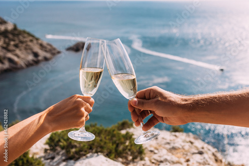 Hands holding champagne glasses over the sea. Romantic vacation. Two hands holding champagne glasses on the background of the sea. A couple in love drinks champagne on the seashore. Copy space © MISHA