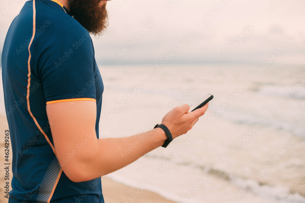 No face photo of a bearded strong man standing on the beach and looking at the phone.