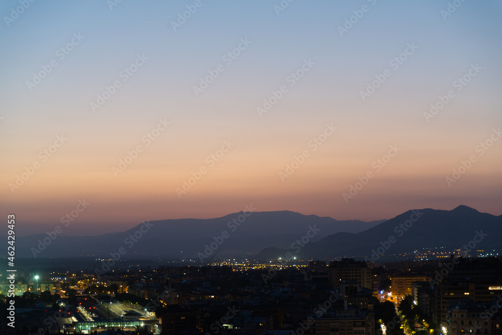 Aerial view of Granada, Andalusia, Spain at night. Urban city light in the foreground. Sierra Nevada mountain range with colorful sunset sky on the background.